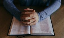 Man's hands clasped in prayer on top of a Bible