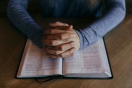 Man's hands clasped in prayer on top of a Bible